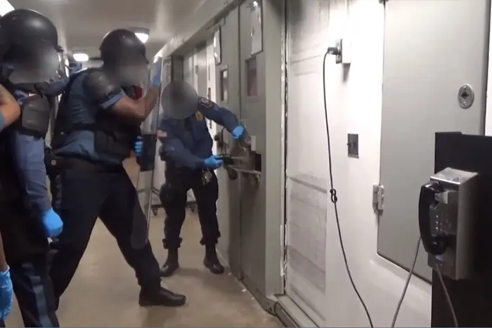 A screenshot of three officers in protective riot gear stand in front of a white door with weapons drawn. Their faces are blurred.